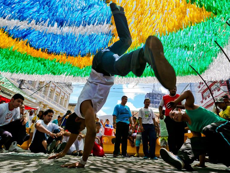 Unter einer brasilianischen Flagge tanzen zwei junge Männer Capoeira: Der eine steht gerade auf seinen Händen, der andere dreht sich am Boden um sich selbst.