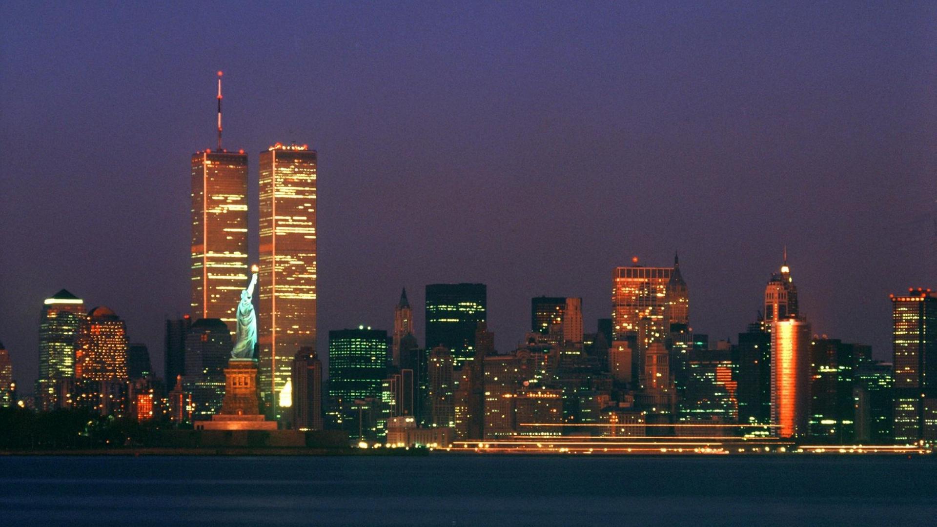 Blick auf die Skyline des New Yorker Stadtteils Manhattan mit den Zwillingstürmen des World Trade Centers und der Freiheitsstatue bei Nacht.
