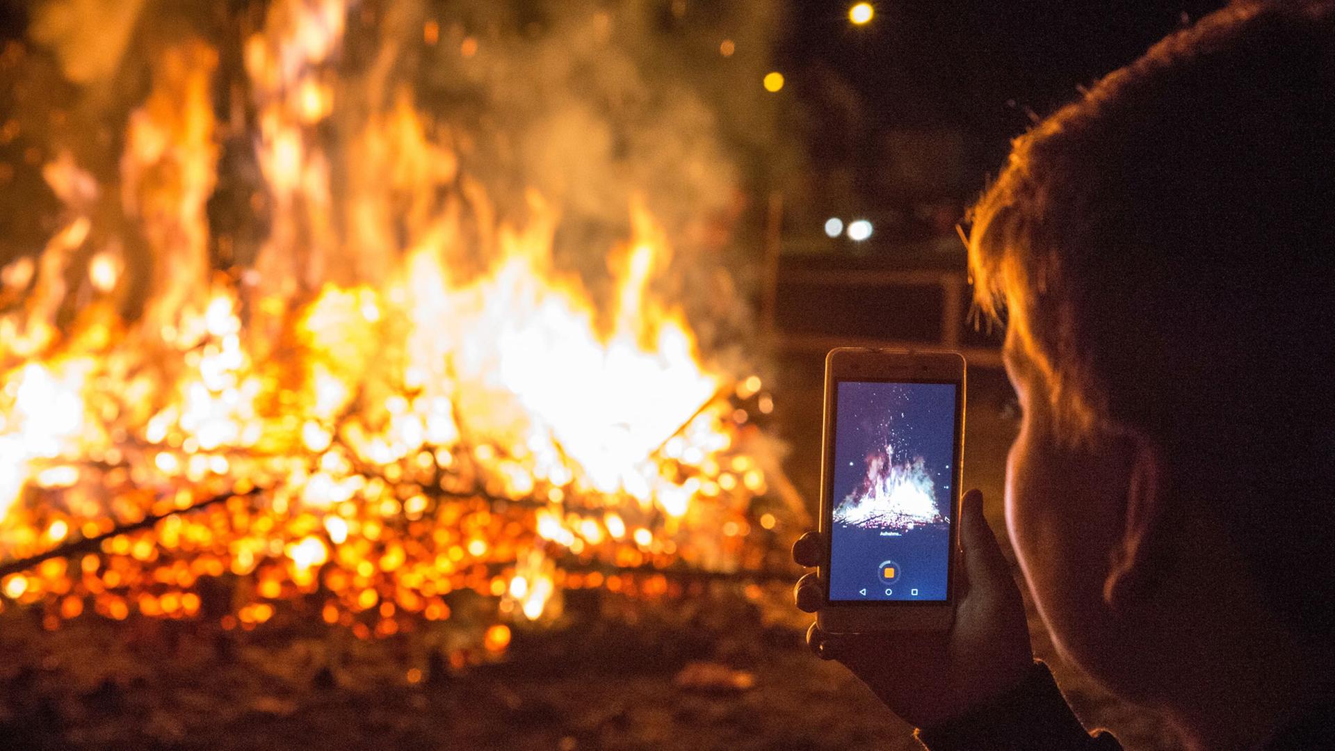 Eine Person filmt mit dem Handy brennende Christbäume auf einem Scheiterhaufen bei Nacht.