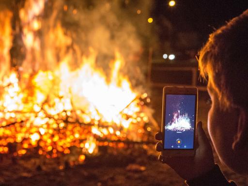 Eine Person filmt mit dem Handy brennende Christbäume auf einem Scheiterhaufen bei Nacht.