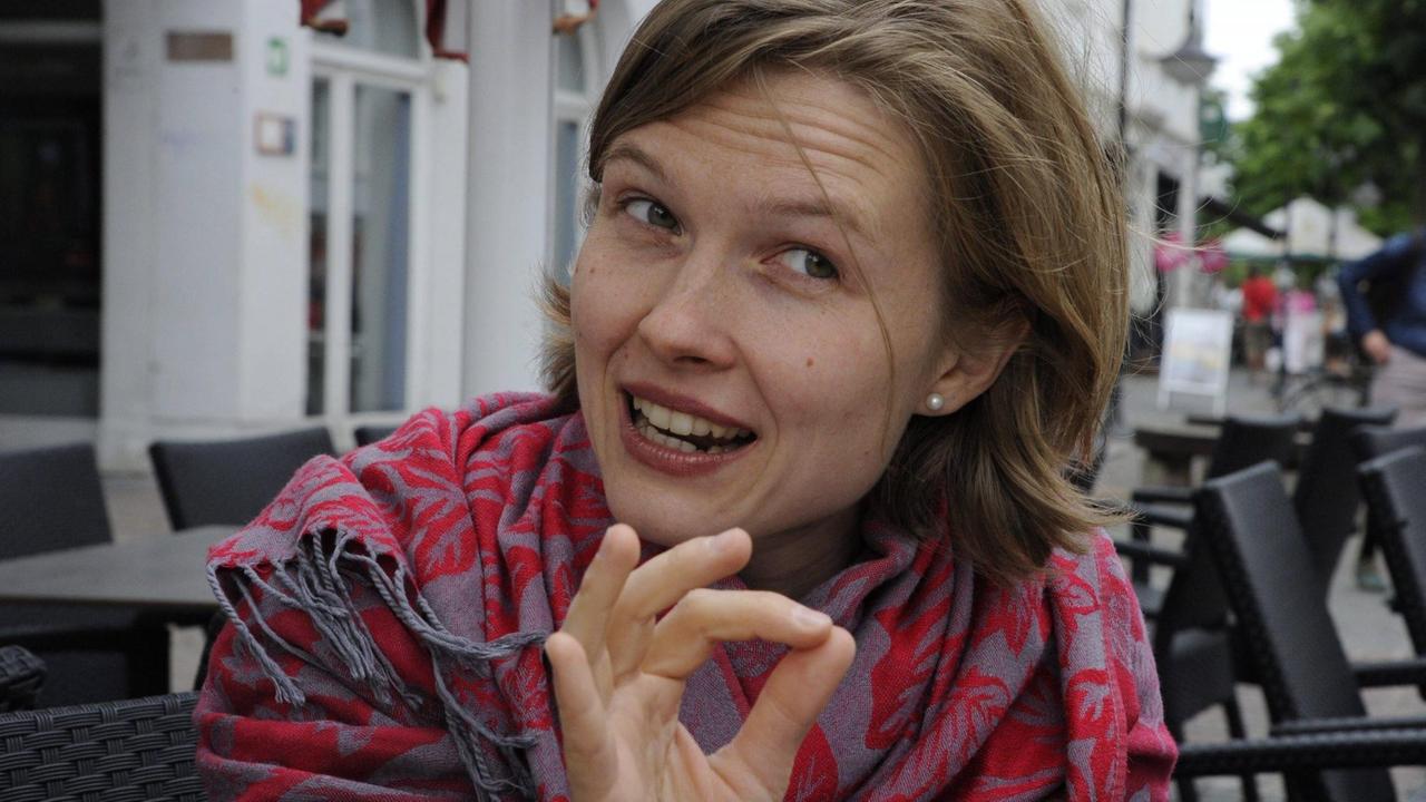 Eine junge Frau mit schulterlangen, blonden Haaren sitzt in einem Straßenkaffee und erzählt, wobei eine ihrer Hände eine Dirigentenbewegung ausführt. Sie hat die Spitzen von Zeigefinger und Damen zu einem Kreiszeichen zusammengeführt. Eine junge Frau mit schulterlangen, blonden Haaren sitzt in einem Straßenkaffee und erzählt, wobei eine ihrer Hände eine Dirigentenbewegung ausführt. Sie hat die Spitzen von Zeigefinger und Damen zu einem Kreiszeichen zusammengeführt.