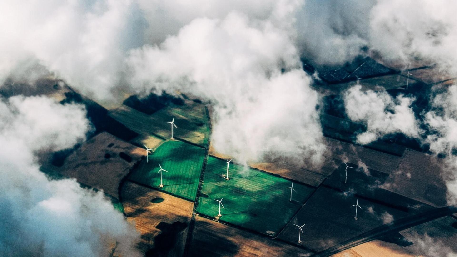 Eine Luftaufnahme zeigt Felder in verschiedenen Grüntönen mit Windkraftanlagen vor einem dramatischen Himmel. Eine Luftaufnahme zeigt Felder in verschiedenen Grüntönen mit Windkraftanlagen vor einem dramatischen Himmel.