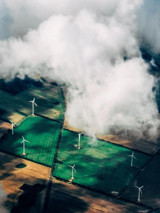 Eine Luftaufnahme zeigt Felder in verschiedenen Grüntönen mit Windkraftanlagen vor einem dramatischen Himmel.
