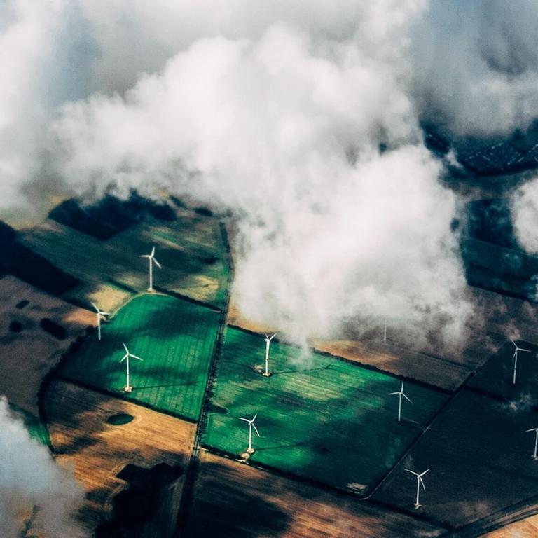 Eine Luftaufnahme zeigt Felder in verschiedenen Grüntönen mit Windkraftanlagen zwischen einem dramatischem Himmel.