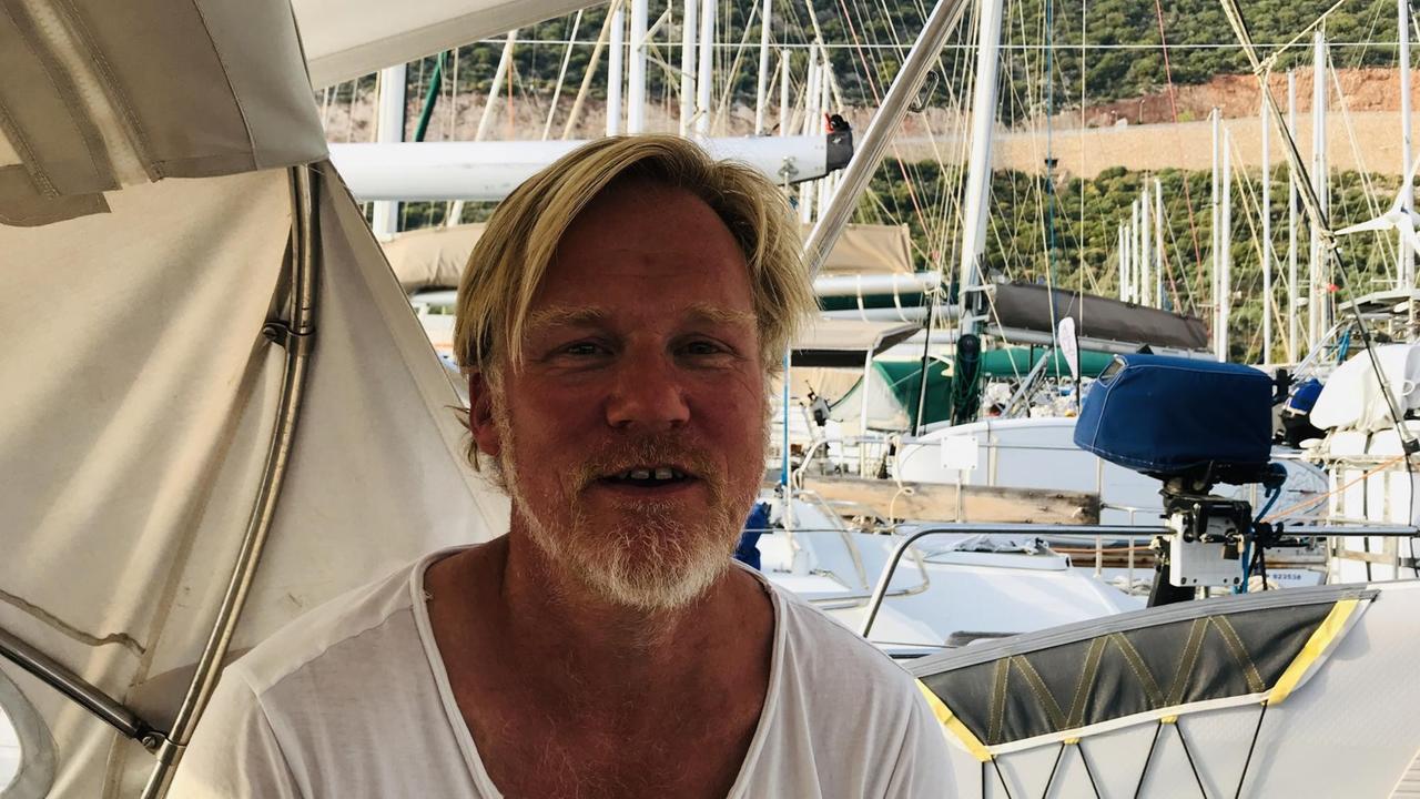 Ein blonder braungebrannter Mann mit Stoppelbart in weißem T-Shirt sitzt auf einem Boot. Im Hintergrund der Hafen. Ein blonder braungebrannter Mann mit Stoppelbart in weißem T-Shirt sitzt auf einem Boot. Im Hintergrund der Hafen.