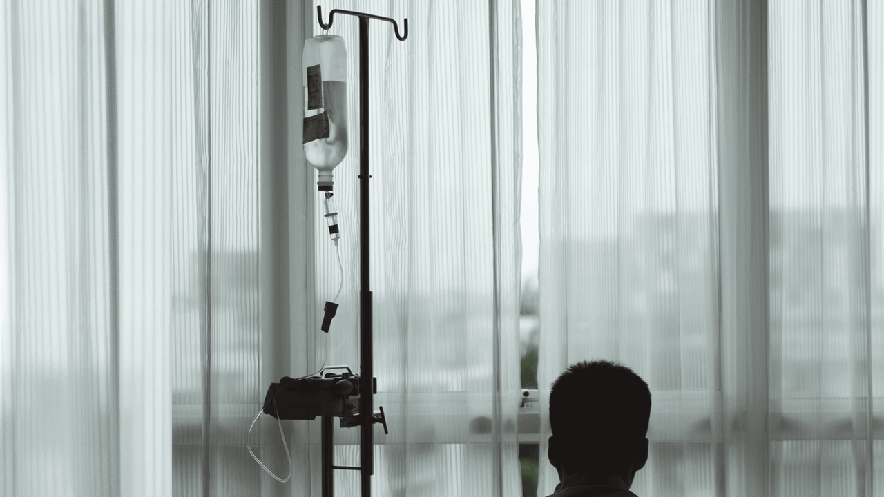 Im Krankenhaus, auf sich gestellt. Ein Mann in einem Krankenhauszimmer schaut durch die Gardine durch das Fenster hinaus.