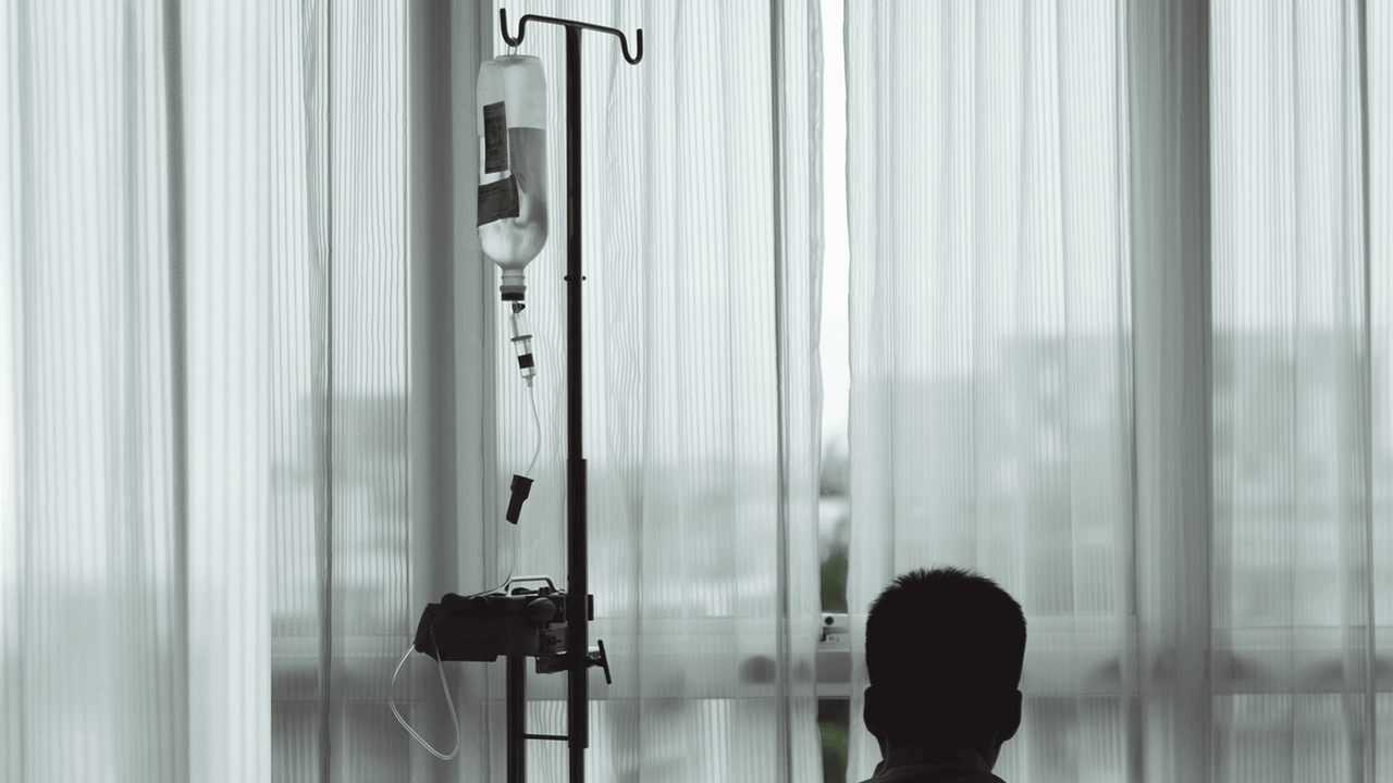 Im Krankenhaus, auf sich gestellt. Ein Mann in einem Krankenhauszimmer schaut durch die Gardine durch das Fenster hinaus. Im Krankenhaus, auf sich gestellt. Ein Mann in einem Krankenhauszimmer schaut durch die Gardine durch das Fenster hinaus.