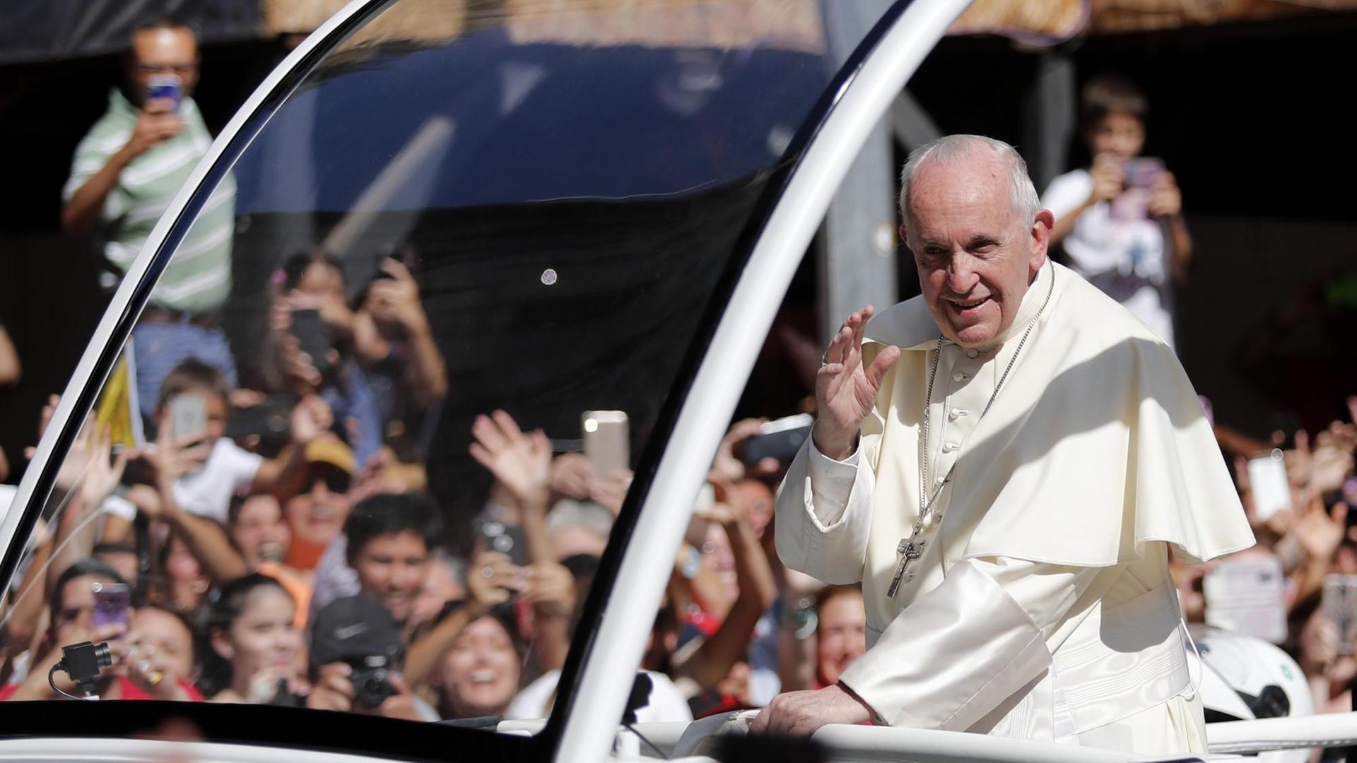 Man sieht den Papst im weißen Gewand lächelnd im Vordergrund, im Hintergrund unscharf jubelnde Zuschauer, die mit Handys fotografieren. Man sieht den Papst im weißen Gewand lächelnd im Vordergrund, im Hintergrund unscharf jubelnde Zuschauer, die mit Handys fotografieren.
