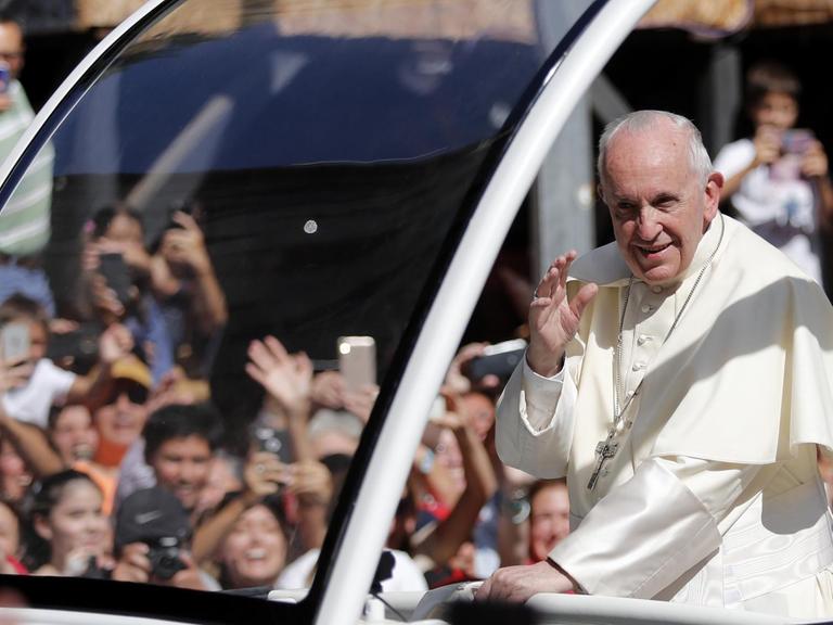 Man sieht den Papst im weißen Gewand lächelnd im Vordergrund, im Hintergrund unscharf jubelnde Zuschauer, die mit Handys fotografieren.