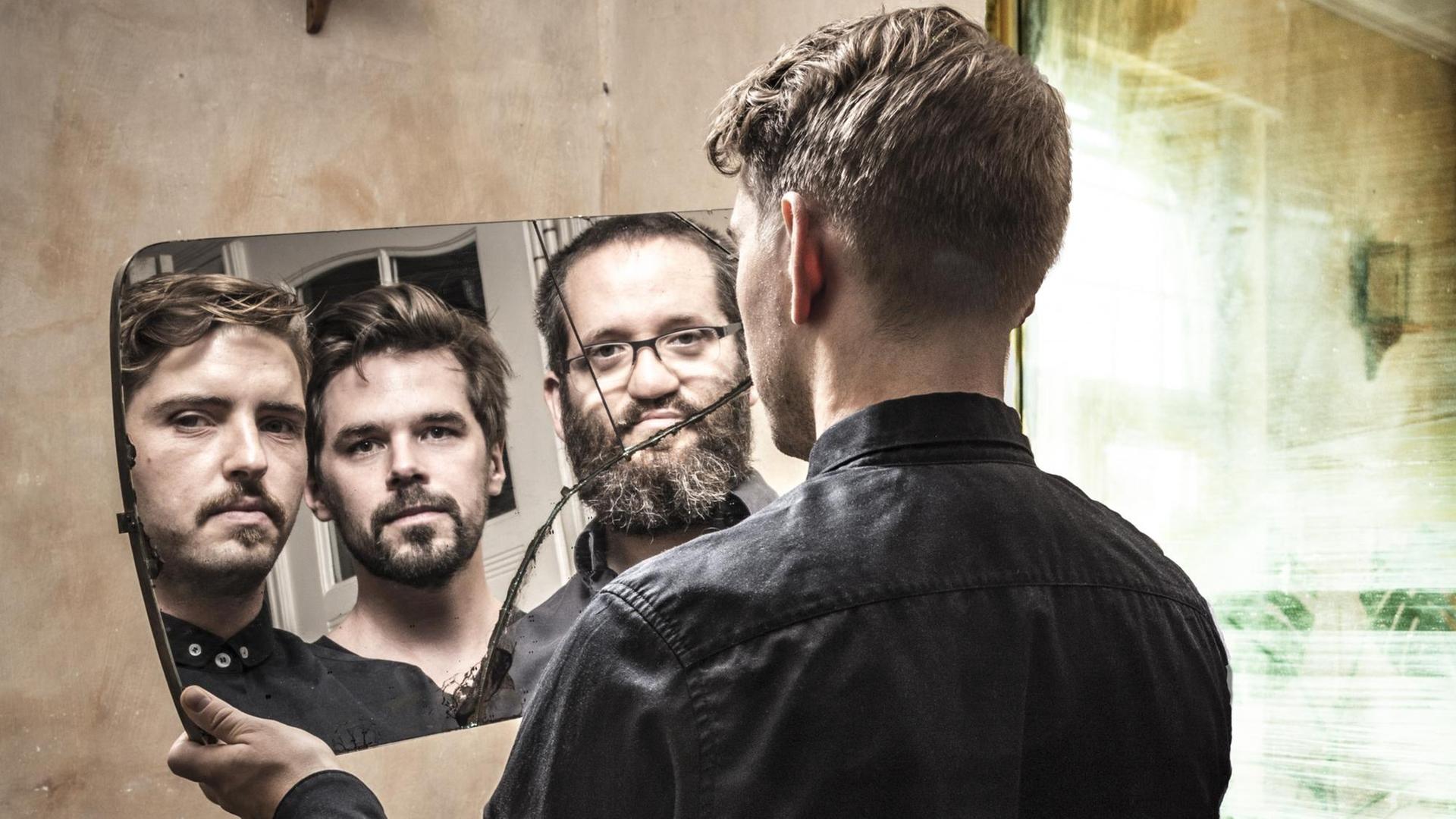 Ein Mann schaut in einen zerbrochenen Spiegel, in dem sich außer ihm zwei weitere Personen widerspiegeln
