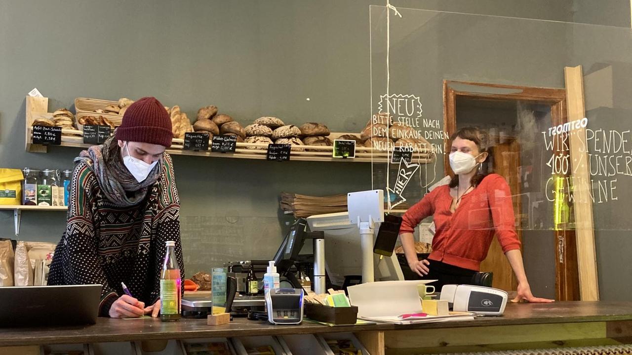 Hinter einem Ladentresen aus Holz stehen ein junger Mann und eine junge Frau, beide tragen FFP2-Masken. Hinter einem Ladentresen aus Holz stehen ein junger Mann und eine junge Frau, beide tragen FFP2-Masken.