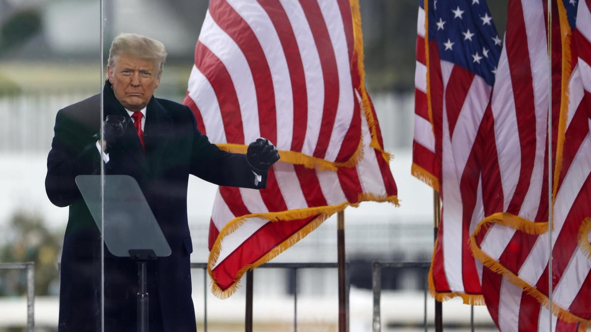 Trump steht hinter Glasscheiben vor der amerikanischen Flagge und ballt seine Hände zu Fäusten. Trump steht hinter Glasscheiben vor der amerikanischen Flagge und ballt seine Hände zu Fäusten.