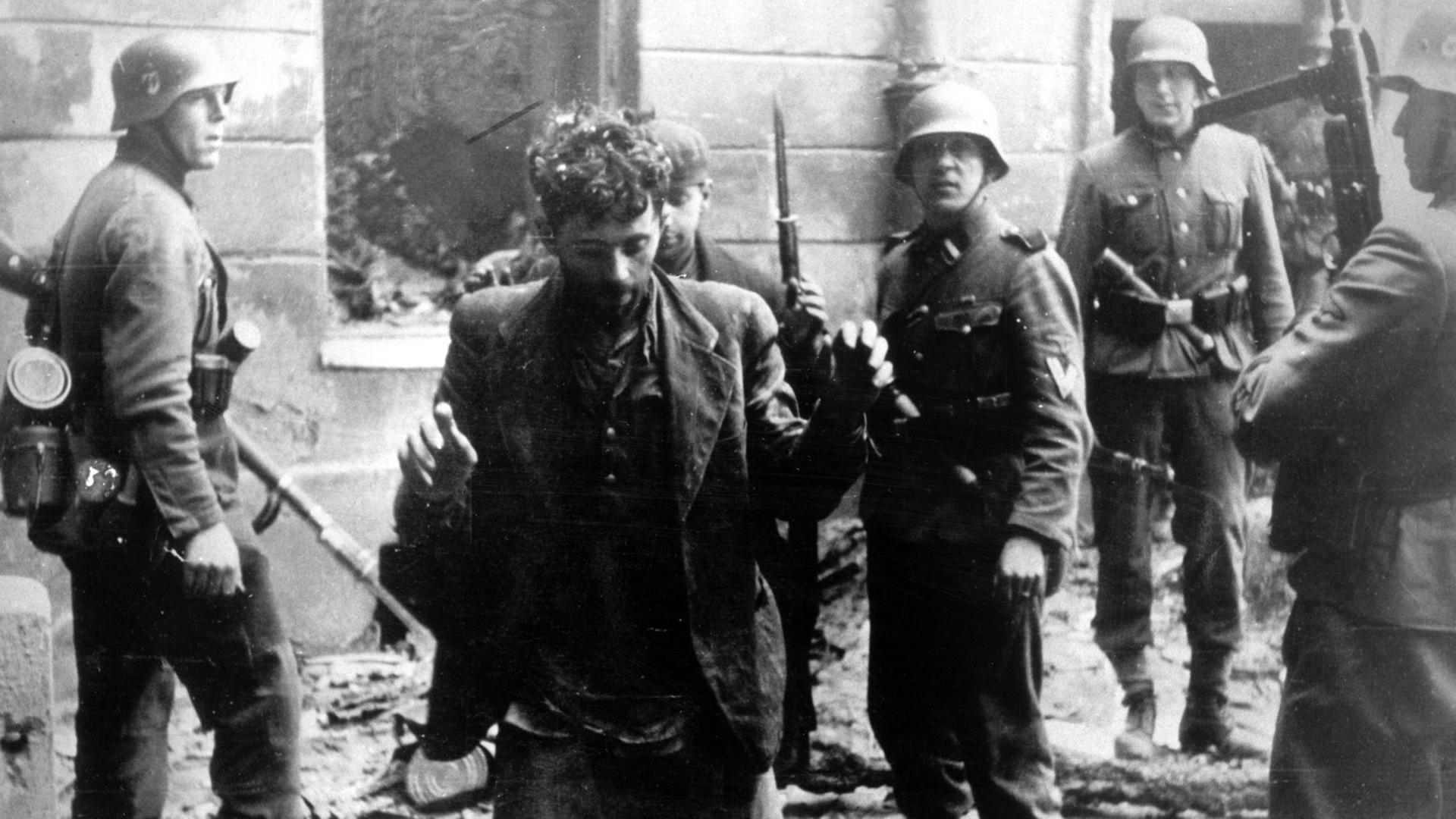 Zwei Juden, die sich in einem Haus versteckt hatten, werden von SS-Soldaten gefangen genommen. Die Aufnahme entstand während des Warschauer Ghetto-Aufstands, der vom 19. April 1943 bis zu seiner blutigen Niederschlagung am 16. Mai 1943 dauerte. Die Nationalsozialisten hatten ein Jahr nach der Besetzung Polens im November 1940 in Warschau ein Ghetto errichtet und dorthin annähernd eine halbe Million Juden verschleppt. Zwischen Juli und September 1942 wurden 300 000 Opfer in den Todeslagern, die meisten in Treblinka, ermordet. Als am 19. April 1943 die SS mit der Verschleppung der restlichen 60 000 Ghetto-Einwohner begann, leisteten mehrere hundert militärisch organisierte Juden bewaffneten Widerstand. Bei den Kämpfen wurden etwa 14 000 jüdische Aufständische getötet. Zwei Juden, die sich in einem Haus versteckt hatten, werden von SS-Soldaten gefangen genommen. Die Aufnahme entstand während des Warschauer Ghetto-Aufstands, der vom 19. April 1943 bis zu seiner blutigen Niederschlagung am 16. Mai 1943 dauerte. Die Nationalsozialisten hatten ein Jahr nach der Besetzung Polens im November 1940 in Warschau ein Ghetto errichtet und dorthin annähernd eine halbe Million Juden verschleppt. Zwischen Juli und September 1942 wurden 300 000 Opfer in den Todeslagern, die meisten in Treblinka, ermordet. Als am 19. April 1943 die SS mit der Verschleppung der restlichen 60 000 Ghetto-Einwohner begann, leisteten mehrere hundert militärisch organisierte Juden bewaffneten Widerstand. Bei den Kämpfen wurden etwa 14 000 jüdische Aufständische getötet.