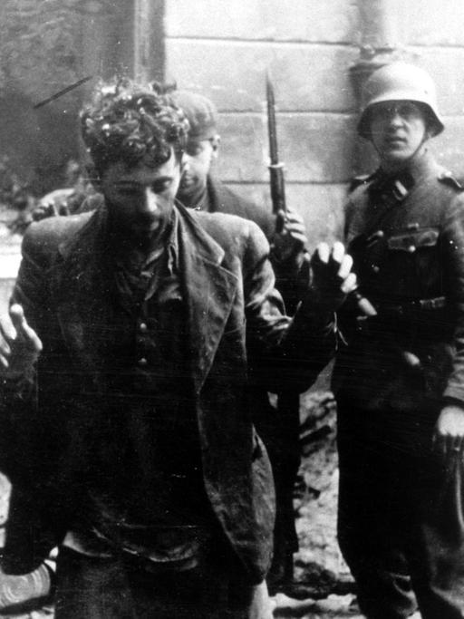 Zwei Juden, die sich in einem Haus versteckt hatten, werden von SS-Soldaten gefangen genommen. Die Aufnahme entstand während des Warschauer Ghetto-Aufstands, der vom 19. April 1943 bis zu seiner blutigen Niederschlagung am 16. Mai 1943 dauerte. Die Nationalsozialisten hatten ein Jahr nach der Besetzung Polens im November 1940 in Warschau ein Ghetto errichtet und dorthin annähernd eine halbe Million Juden verschleppt. Zwischen Juli und September 1942 wurden 300 000 Opfer in den Todeslagern, die meisten in Treblinka, ermordet. Als am 19. April 1943 die SS mit der Verschleppung der restlichen 60 000 Ghetto-Einwohner begann, leisteten mehrere hundert militärisch organisierte Juden bewaffneten Widerstand. Bei den Kämpfen wurden etwa 14 000 jüdische Aufständische getötet.