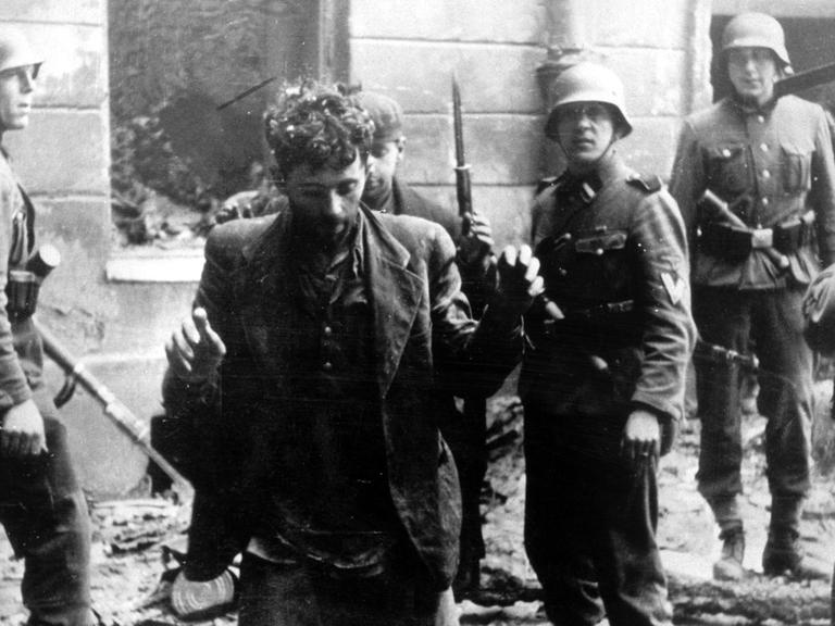 Zwei Juden, die sich in einem Haus versteckt hatten, werden von SS-Soldaten gefangen genommen. Die Aufnahme entstand während des Warschauer Ghetto-Aufstands, der vom 19. April 1943 bis zu seiner blutigen Niederschlagung am 16. Mai 1943 dauerte. Die Nationalsozialisten hatten ein Jahr nach der Besetzung Polens im November 1940 in Warschau ein Ghetto errichtet und dorthin annähernd eine halbe Million Juden verschleppt. Zwischen Juli und September 1942 wurden 300 000 Opfer in den Todeslagern, die meisten in Treblinka, ermordet. Als am 19. April 1943 die SS mit der Verschleppung der restlichen 60 000 Ghetto-Einwohner begann, leisteten mehrere hundert militärisch organisierte Juden bewaffneten Widerstand. Bei den Kämpfen wurden etwa 14 000 jüdische Aufständische getötet.