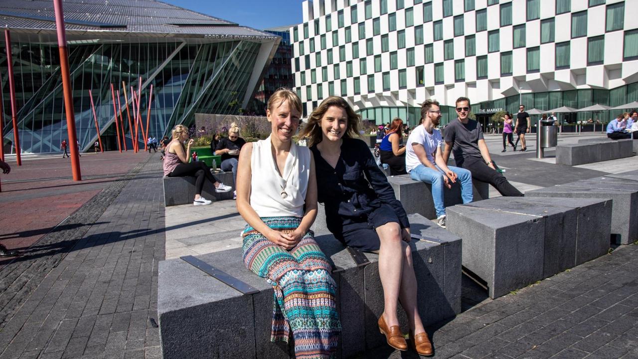 Zwei Frauen sitzen bei Sonnenschein auf einem Steinblock in der Mittagspause vor Bürogebäuden aus Glas, Stahl und Beton. Zwei Frauen sitzen bei Sonnenschein auf einem Steinblock in der Mittagspause vor Bürogebäuden aus Glas, Stahl und Beton.
