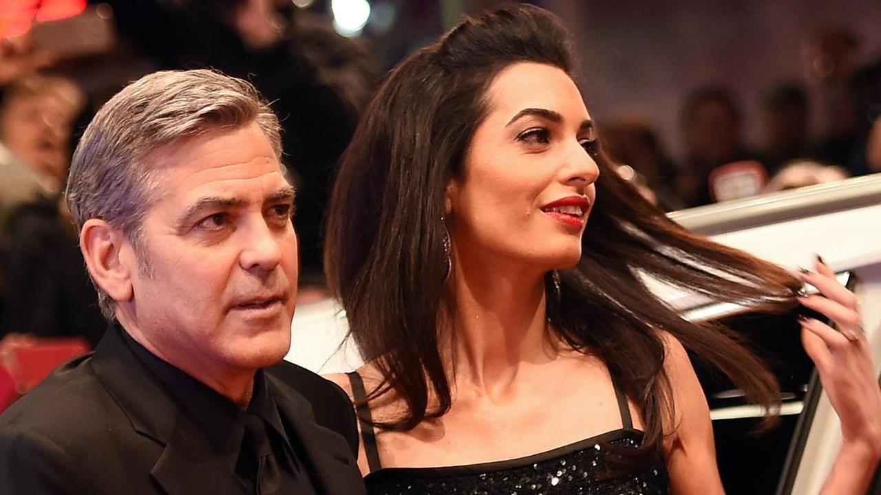 Clooney und Amal schauen engumschlungen in die Kameras, Amal zieht an ihren Haaren. Er trägt einen schwarzen Anzug, sie ein schwarzes schulterfreies Kleid. Clooney und Amal schauen engumschlungen in die Kameras, Amal zieht an ihren Haaren. Er trägt einen schwarzen Anzug, sie ein schwarzes schulterfreies Kleid.