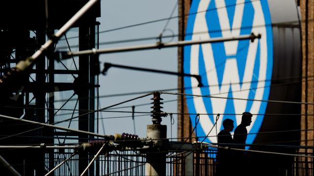 Die Silhouetten von zwei Männern auf einer Brücke zeichnen sich am 10.05.2016 vor dem großen Volkswagen-Logo am Kraftwerk am VW-Werk in Wolfsburg (Niedersachsen) ab.