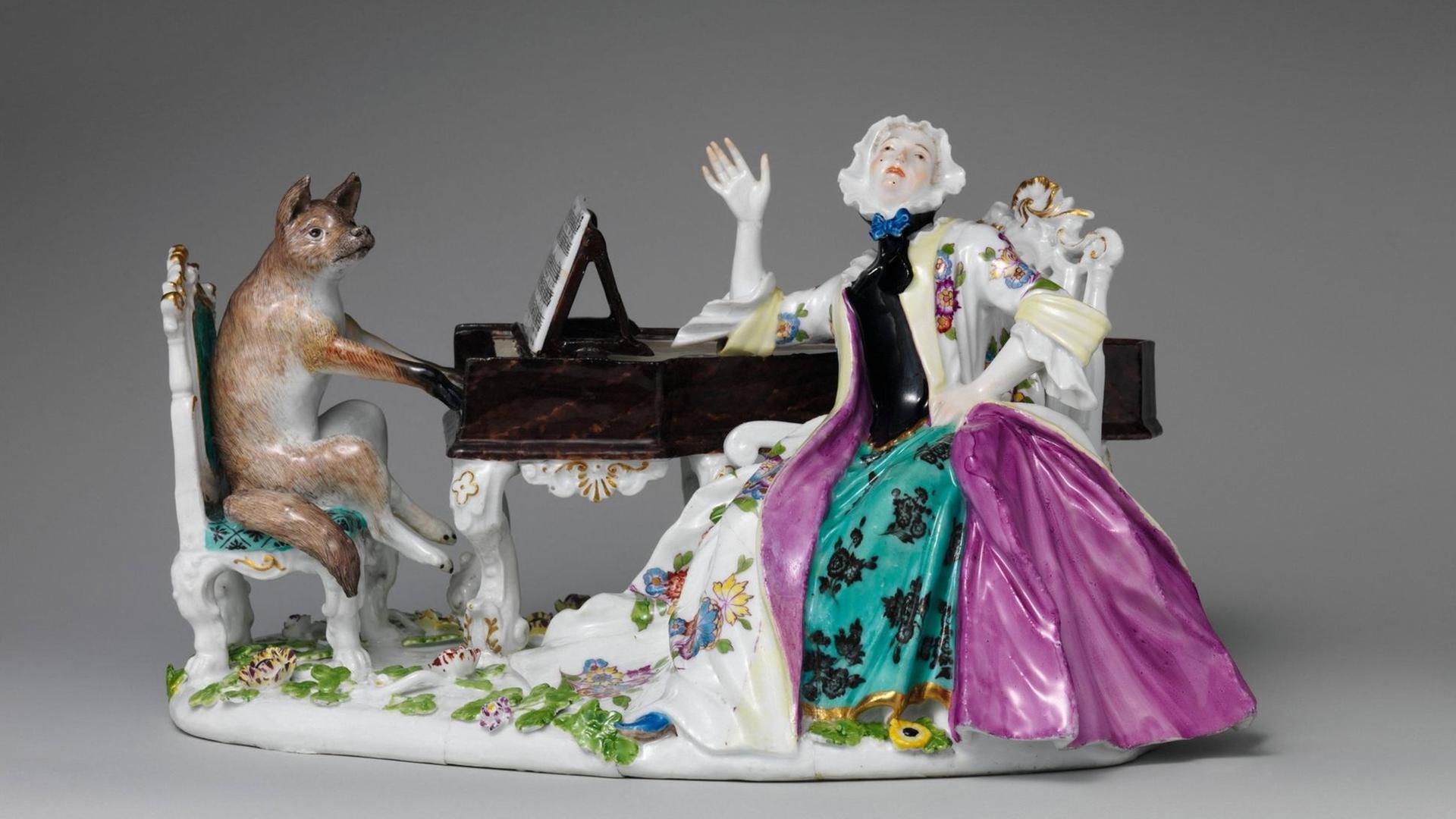 Eine Figurengruppe aus Porzellan zeigt eine barock gekleidete Frau neben einem Flügel sitzend, das Instrument wird von einem Fuchs gespielt. Eine Figurengruppe aus Porzellan zeigt eine barock gekleidete Frau neben einem Flügel sitzend, das Instrument wird von einem Fuchs gespielt.
