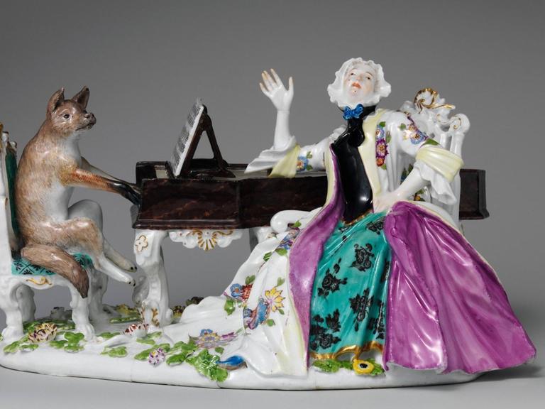 Eine Figurengruppe aus Porzellan zeigt eine barock gekleidete Frau neben einem Flügel sitzend, das Instrument wird von einem Fuchs gespielt.