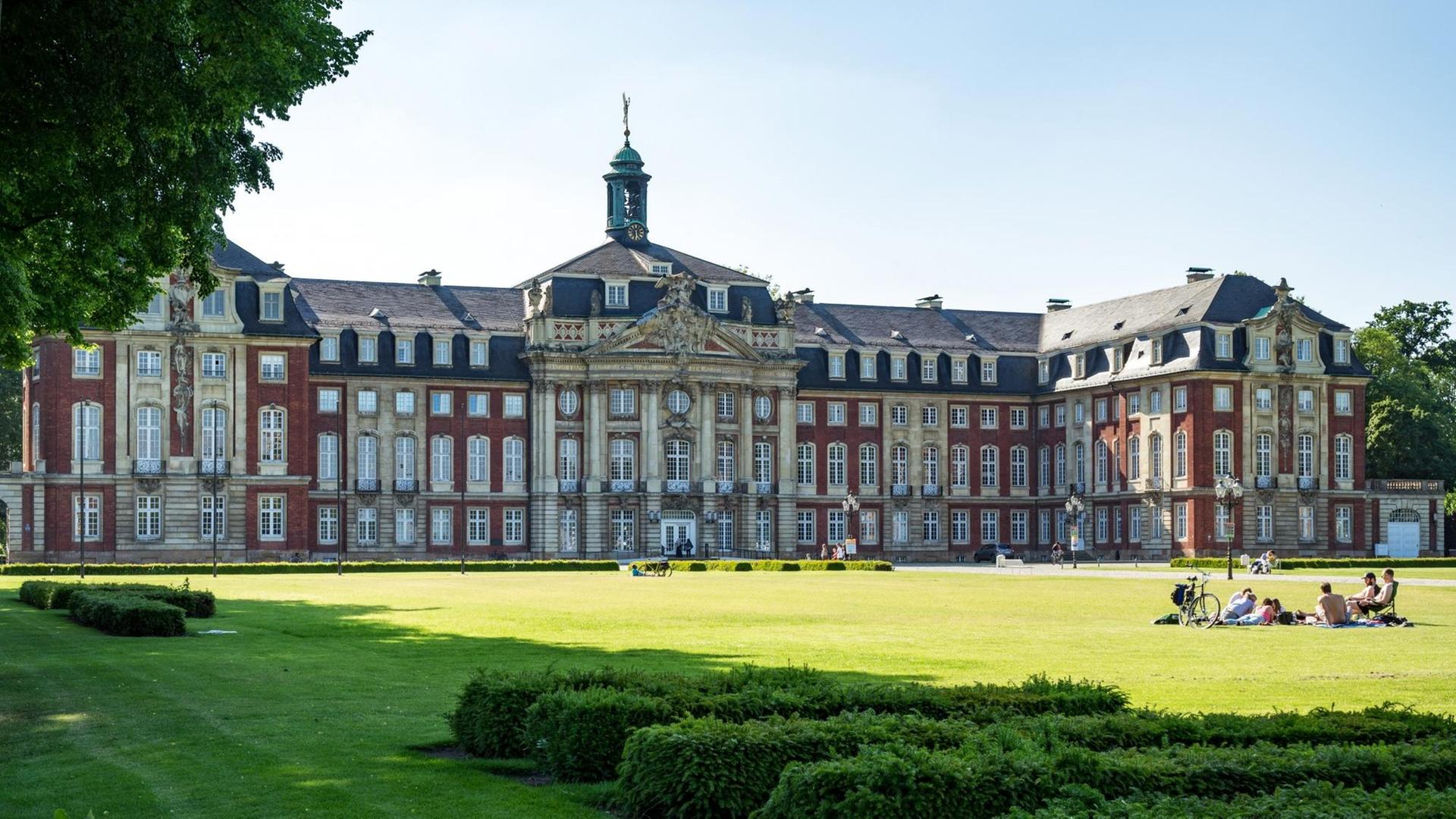 Rotes Schloss, im Vordergrund eine grüne Wiese, auf der eine kleine Gruppe Menschen sitzt. Rotes Schloss, im Vordergrund eine grüne Wiese, auf der eine kleine Gruppe Menschen sitzt.