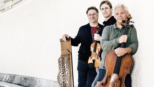 Der Geiger David McCarroll, der Cellist Clemens Hagen und der Pianist Stefan Mendl vom Wiener Klaviertrio stehen an eine weiße Wand gelehnt.