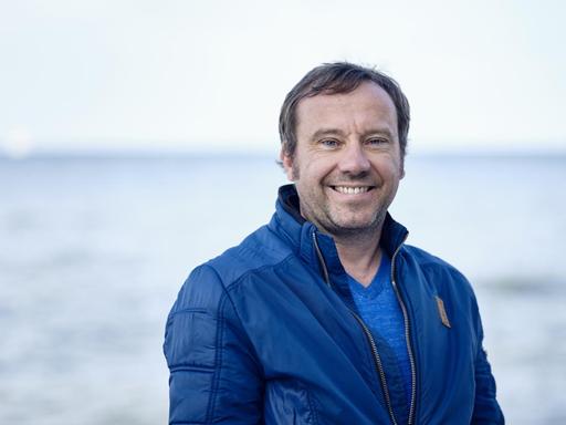 Gregor Sander steht am Meer und lächelt in die Kamera.