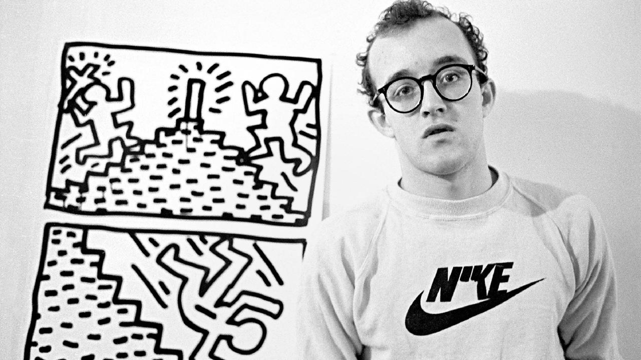 Keith Haring steht vor einer zweiteiligen Bilderserie aus dem Jahr 1982. Darauf sind Strichmännchen auf einem Hügel zu erkennen.