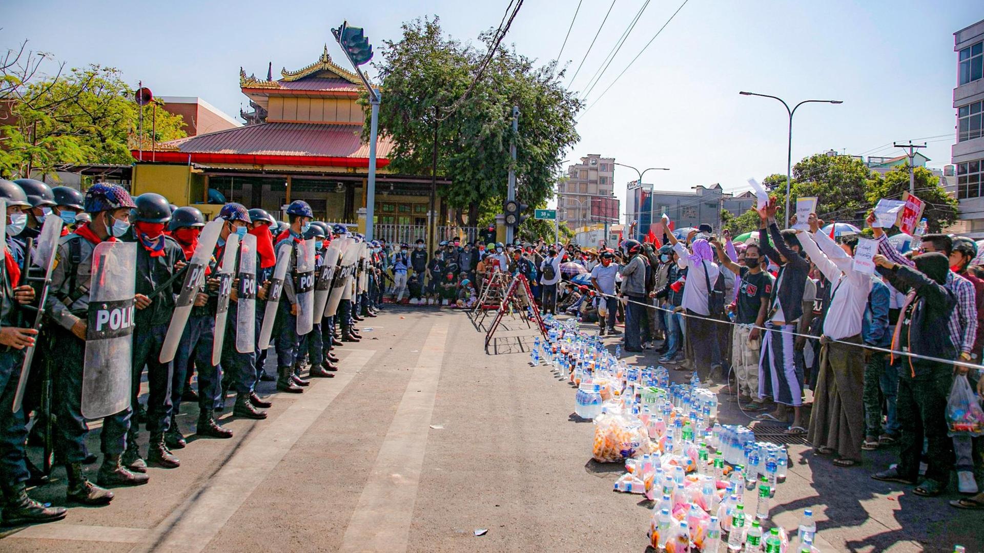 Polizisten und Demonstranten stehen sich an einer Straße in der Stadt Mandalay in Myanmar gegenüber. Die Protestierenden habe Wasserflaschen hinter die Absperrung gestellt und halten Schilder in die Höhe. Polizisten und Demonstranten stehen sich an einer Straße in der Stadt Mandalay in Myanmar gegenüber. Die Protestierenden habe Wasserflaschen hinter die Absperrung gestellt und halten Schilder in die Höhe.