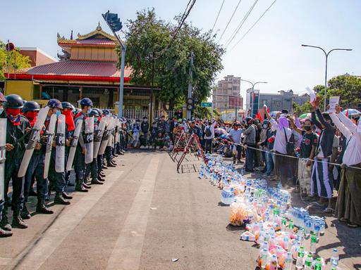 Polizisten und Demonstranten stehen sich an einer Straße in der Stadt Mandalay in Myanmar gegenüber. Die Protestierenden habe Wasserflaschen hinter die Absperrung gestellt und halten Schilder in die Höhe.