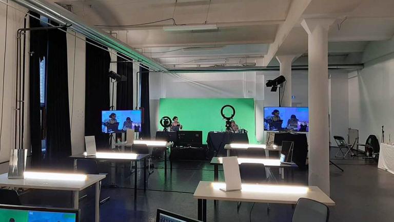 Aufnahme vom digitalen Facefilter-Workshop am Theater Hebbel am Ufer in Berlin. In einem Arbeitsraum mit beleuchtet Tischen, einem Green-Screen und zwei Personen vor der Kamera zu sehen. Aufnahme vom digitalen Facefilter-Workshop am Theater Hebbel am Ufer in Berlin. In einem Arbeitsraum mit beleuchtet Tischen, einem Green-Screen und zwei Personen vor der Kamera zu sehen.