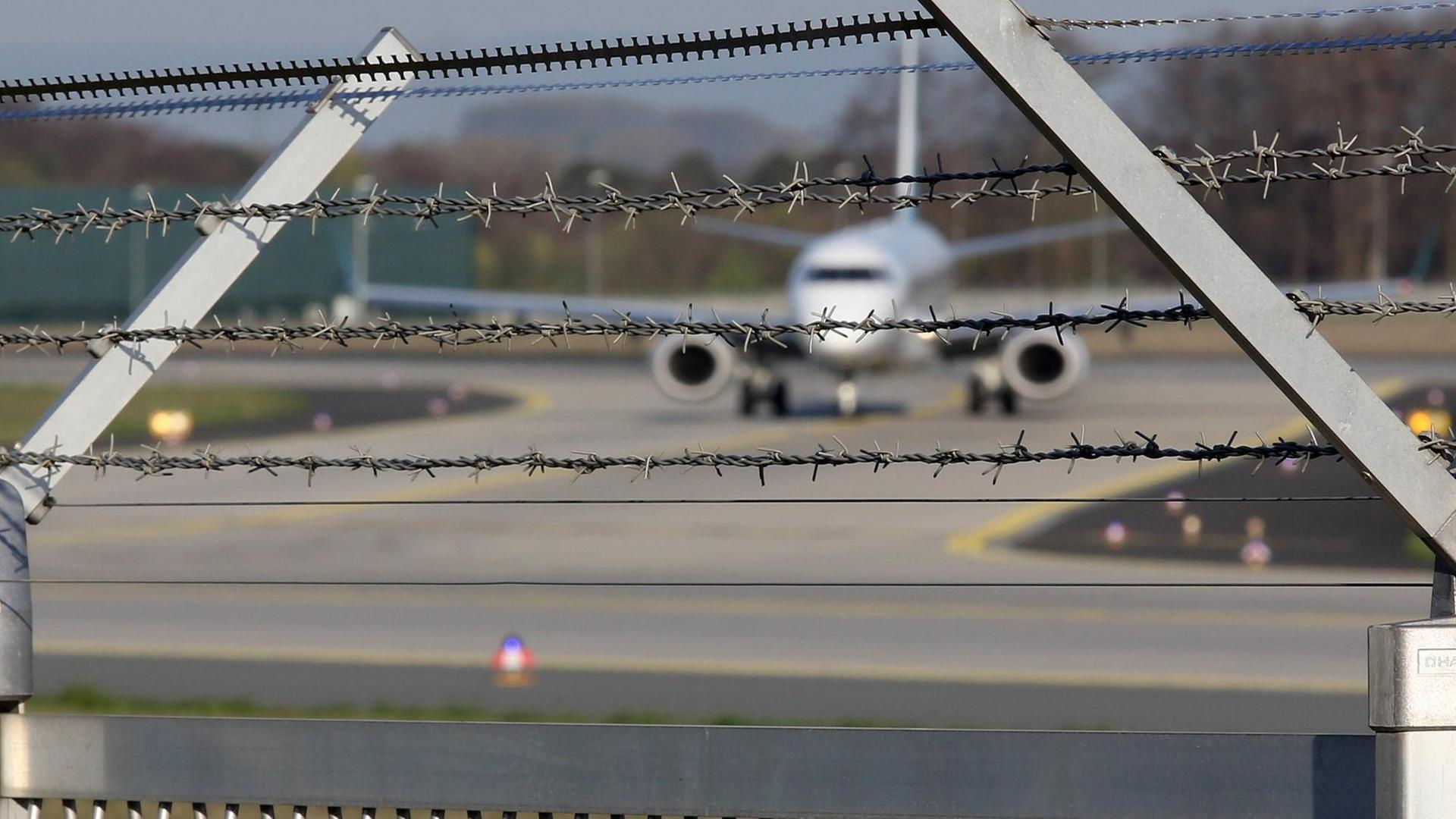 Symbolfoto zum Thema Abschiebung: Im Vordergrund ist ein Zaun mit Sacheldraht zu sehen, im Hintergrund ein Flugzeug auf dem Flughafen Frankfurt am Main. Symbolfoto zum Thema Abschiebung: Im Vordergrund ist ein Zaun mit Sacheldraht zu sehen, im Hintergrund ein Flugzeug auf dem Flughafen Frankfurt am Main.