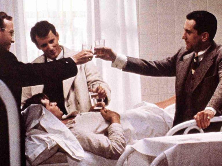 Szene aus dem Film "Es war einmal in Amerika" von 1984 mit den SchauspielernTreat Williams (liegend ), James Woods, Robert De Niro in der Regie von Sergio Leone. Am Krankenbett wird mit Alkohol angestoßen.