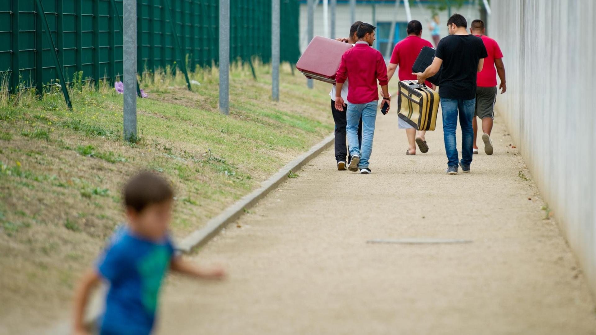 Flüchtlinge laufen am 27.08.2015 mit Koffern bepackt auf einem Weg einer Erstaufnahmeeinrichtung für Flüchtlinge in Ingelheim (Rheinland-Pfalz) entlang, während ein Kind im Vordergrund vorbeiläuft. Flüchtlinge laufen am 27.08.2015 mit Koffern bepackt auf einem Weg einer Erstaufnahmeeinrichtung für Flüchtlinge in Ingelheim (Rheinland-Pfalz) entlang, während ein Kind im Vordergrund vorbeiläuft.