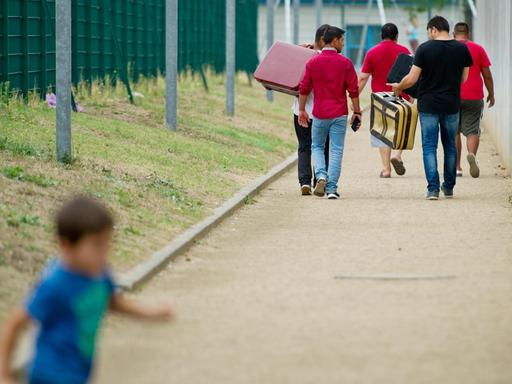 Flüchtlinge laufen am 27.08.2015 mit Koffern bepackt auf einem Weg einer Erstaufnahmeeinrichtung für Flüchtlinge in Ingelheim (Rheinland-Pfalz) entlang, während ein Kind im Vordergrund vorbeiläuft.