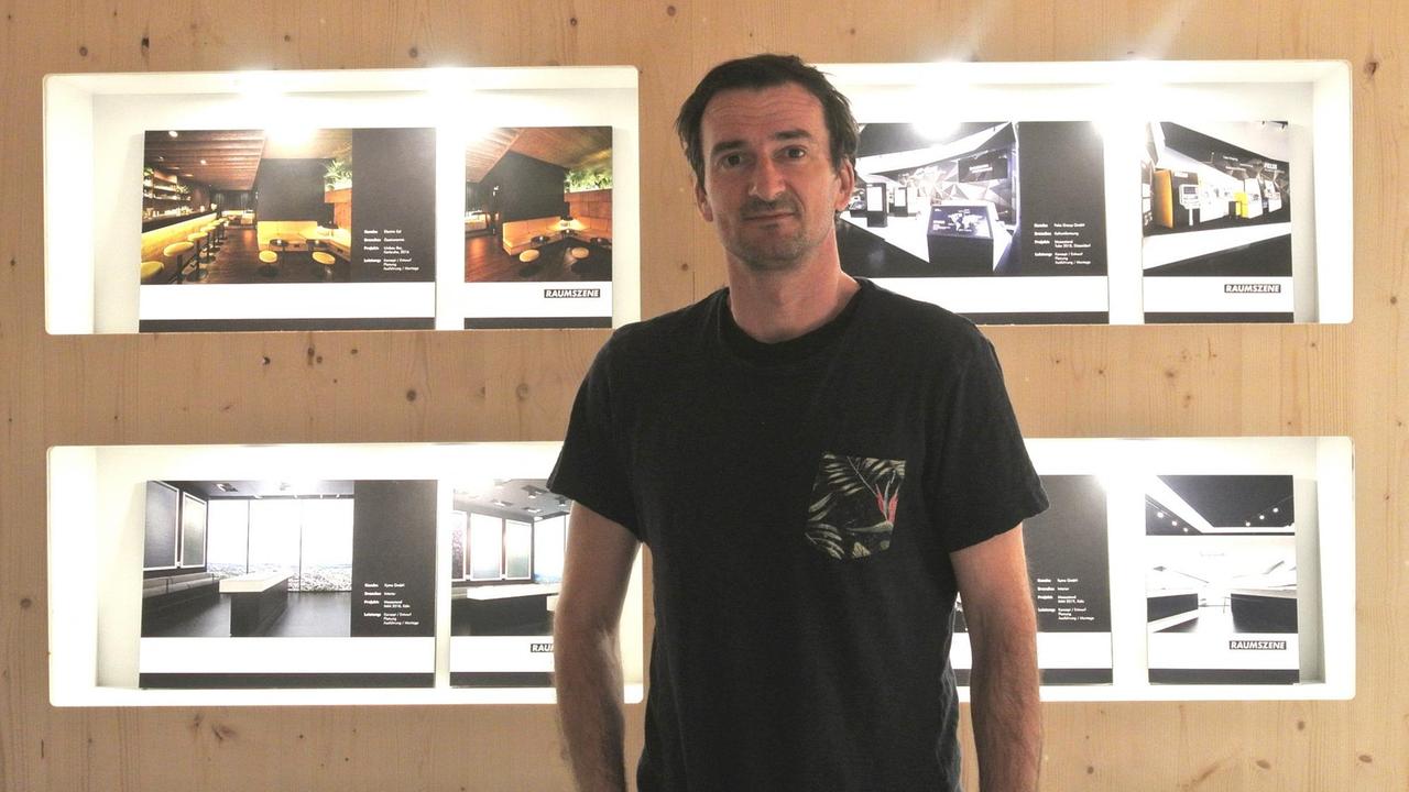 Ein Mann mit T-Shirt steht vor einer Holzwand, auf der Blätter mit Bauentwürfen befestigt sind. Ein Mann mit T-Shirt steht vor einer Holzwand, auf der Blätter mit Bauentwürfen befestigt sind.