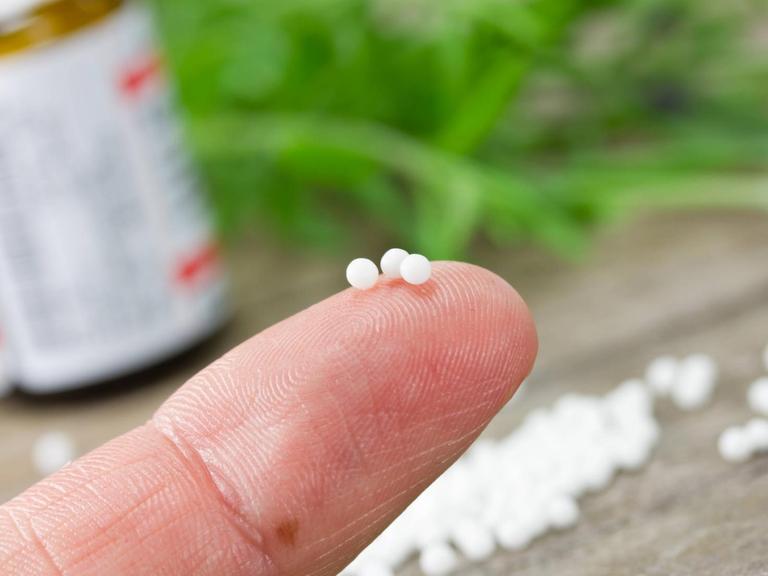 Auf einer Fingerspitze liegen drei weiße Kügelchen, im Hintergrund liegen weitere Kügelchen auf einer Holzplatte, in der linken Ecke ist eine kleine Arzneiflasche zu sehen