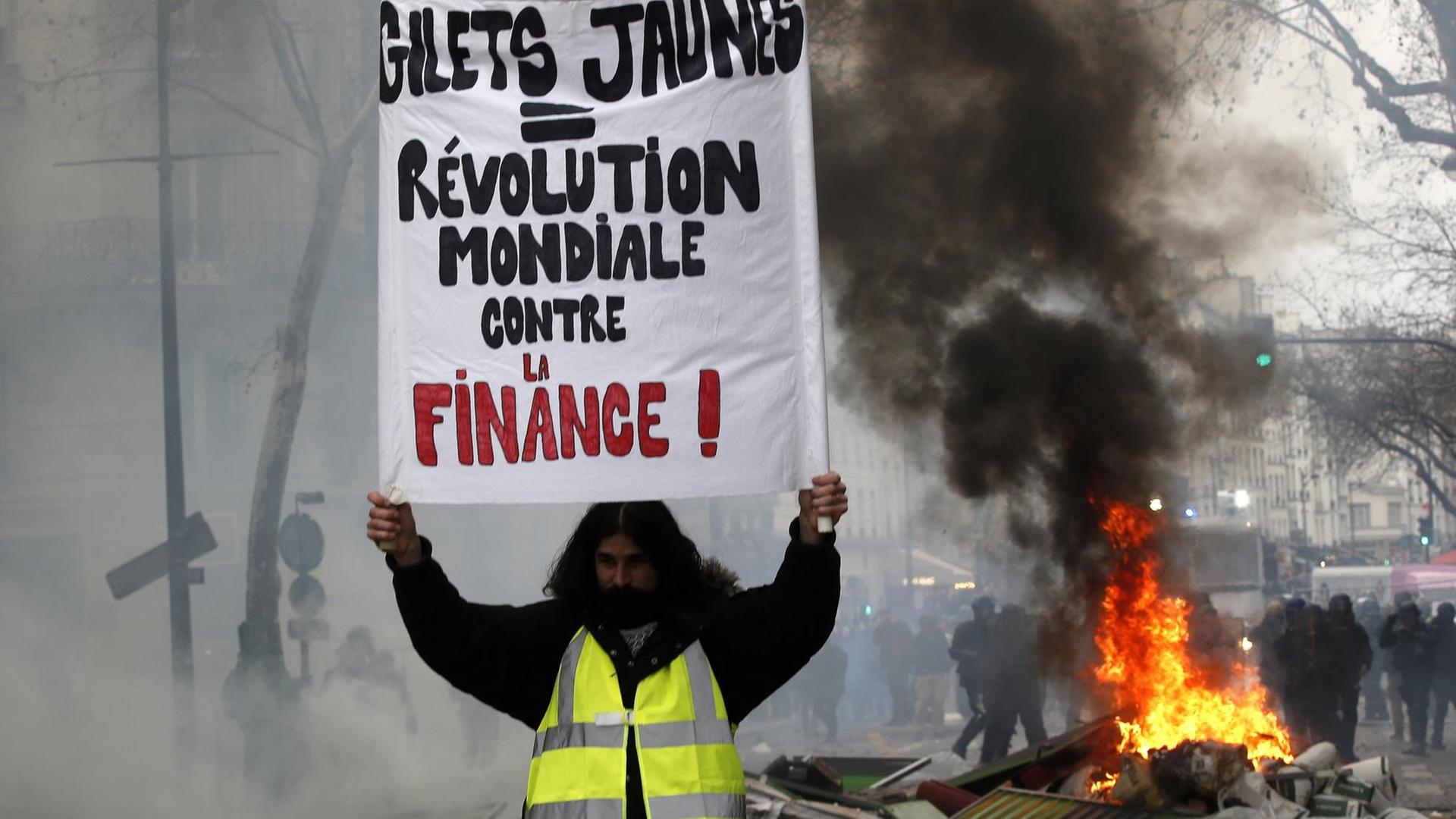 Neue Proteste in Paris. Ein Demonstrant hält ein Schild mit der Aufschrift "Gelbwesten = weltweite Revolution gegen die Finanzwirtschaft". Neue Proteste in Paris. Ein Demonstrant hält ein Schild mit der Aufschrift "Gelbwesten = weltweite Revolution gegen die Finanzwirtschaft".