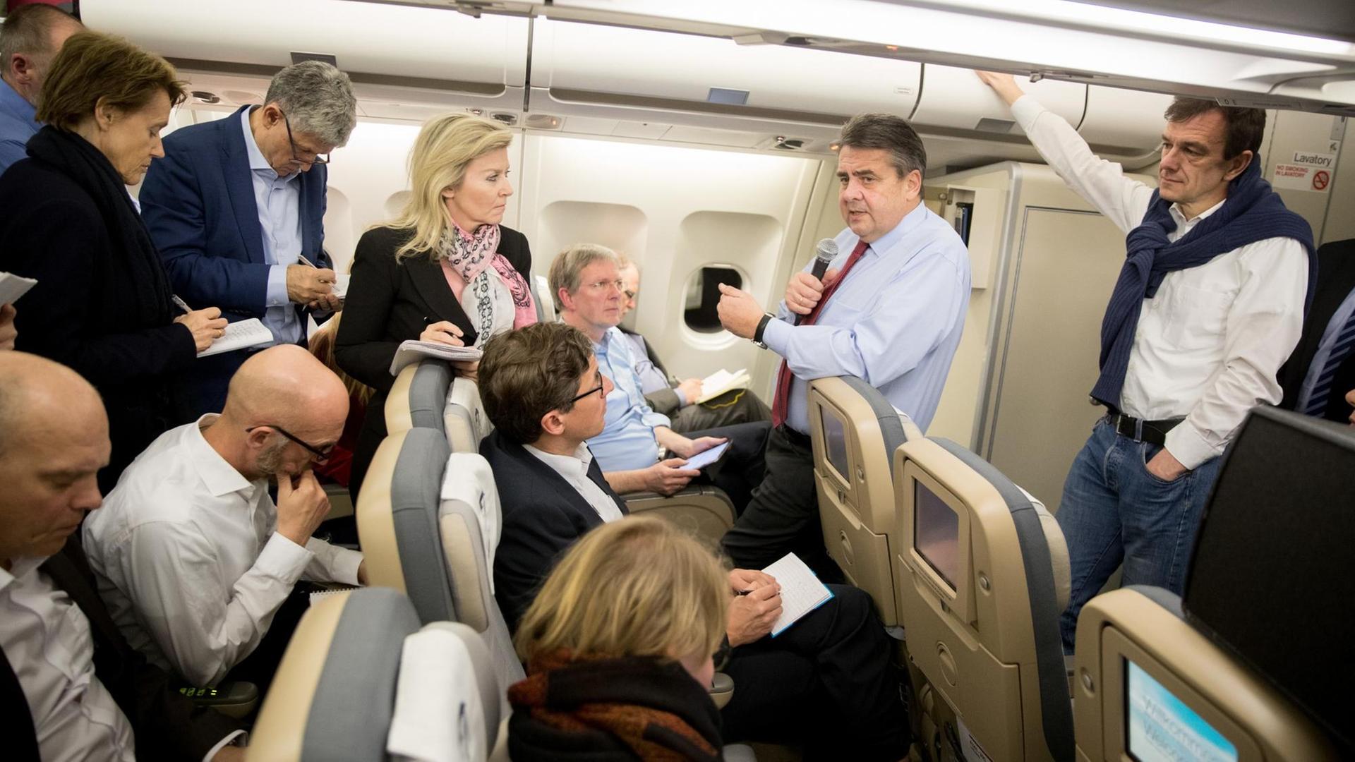 In einem Flugzeug spricht Sigmar Gabriel in ein Mikrofon, um ihn herum stehen und sitzen Journalisten In einem Flugzeug spricht Sigmar Gabriel in ein Mikrofon, um ihn herum stehen und sitzen Journalisten