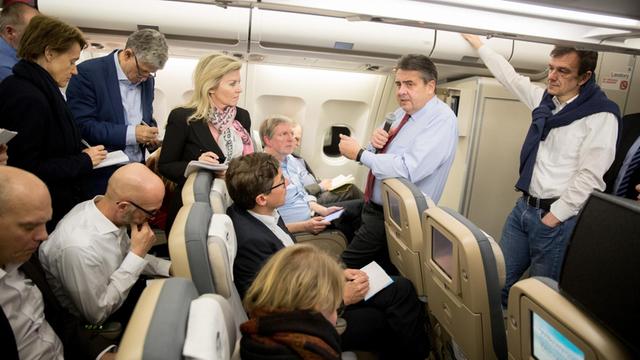 In einem Flugzeug spricht Sigmar Gabriel in ein Mikrofon, um ihn herum stehen und sitzen Journalisten