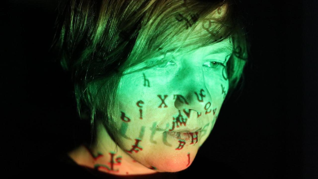 Portrait einer Frau mit schwarzen Haaren. Sie ist grün und orange angestrahlt. Auf dem Gesicht spiegeln sich Buchstaben. Portrait einer Frau mit schwarzen Haaren. Sie ist grün und orange angestrahlt. Auf dem Gesicht spiegeln sich Buchstaben.