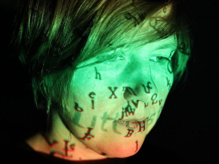Portrait einer Frau mit schwarzen Haaren. Sie ist grün und orange angestrahlt. Auf dem Gesicht spiegeln sich Buchstaben.