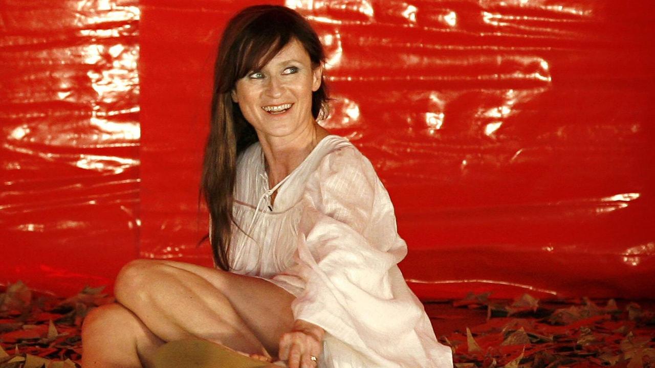 Eine braunhaarige Schauspielerin sitzt lächelnd vor einer grellroter Wand.