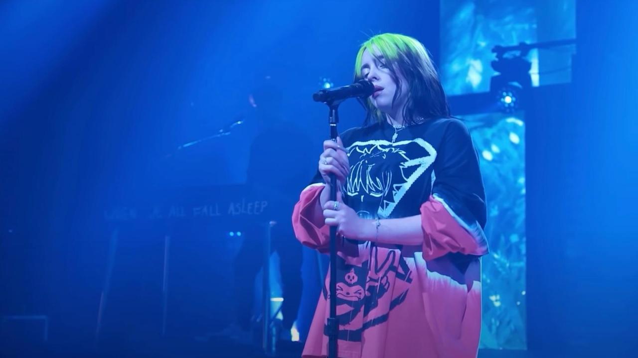 Billie Eilish steht singend auf einer Bühne und umklammert mit den Händen den Sänder des Mikrofons. Sie trägt ein sehr weites, sackartiges und gemustertes Oberteil. Ihre Haare sind schwarz und neon-grün gefärbt. Billie Eilish steht singend auf einer Bühne und umklammert mit den Händen den Sänder des Mikrofons. Sie trägt ein sehr weites, sackartiges und gemustertes Oberteil. Ihre Haare sind schwarz und neon-grün gefärbt.