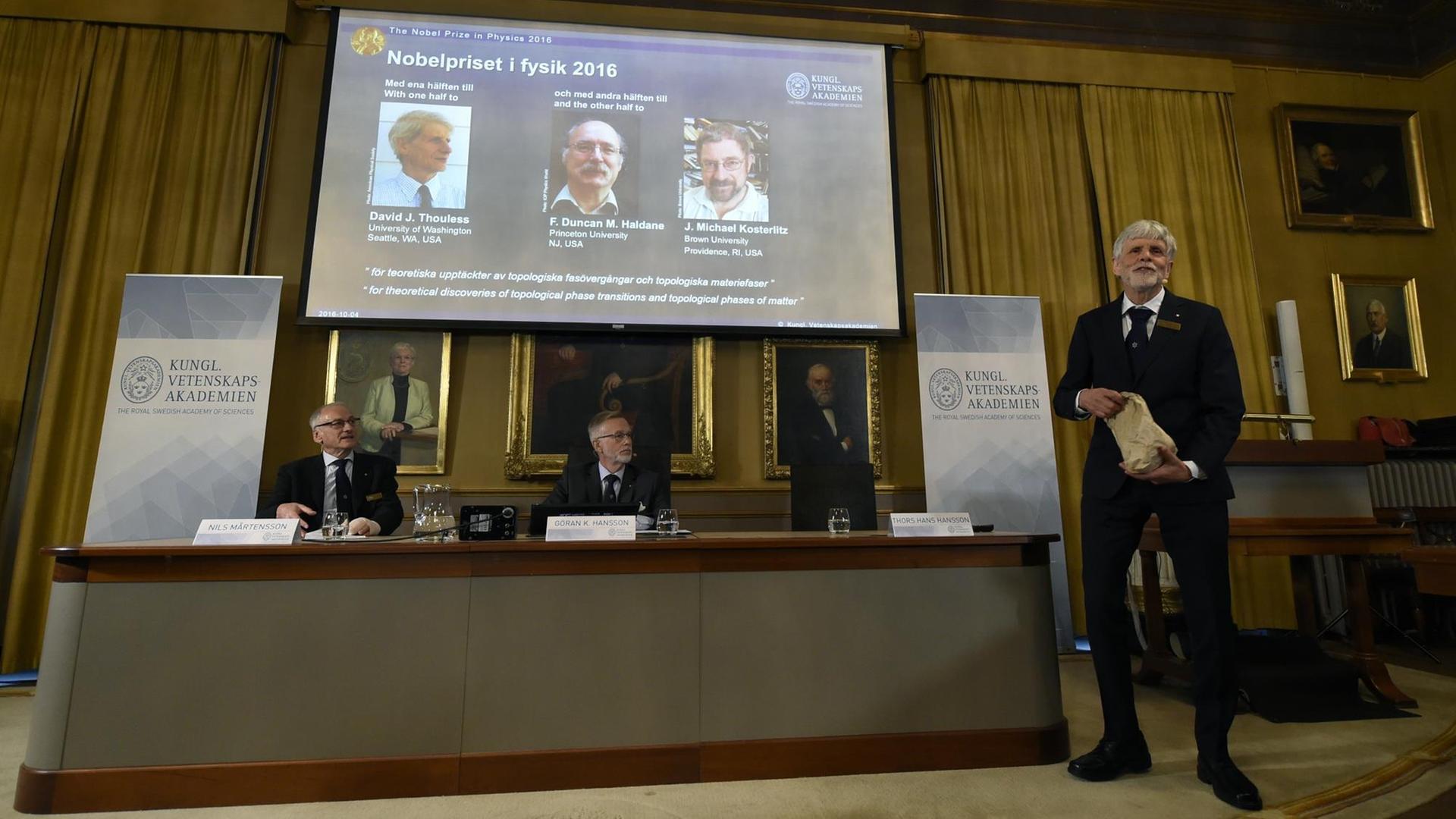 Während der Pressekonferenz in Stockholm werden die Porträts der drei Preisträger Thouless, Haldane und Kosterlitz eingeblendet Während der Pressekonferenz in Stockholm werden die Porträts der drei Preisträger Thouless, Haldane und Kosterlitz eingeblendet