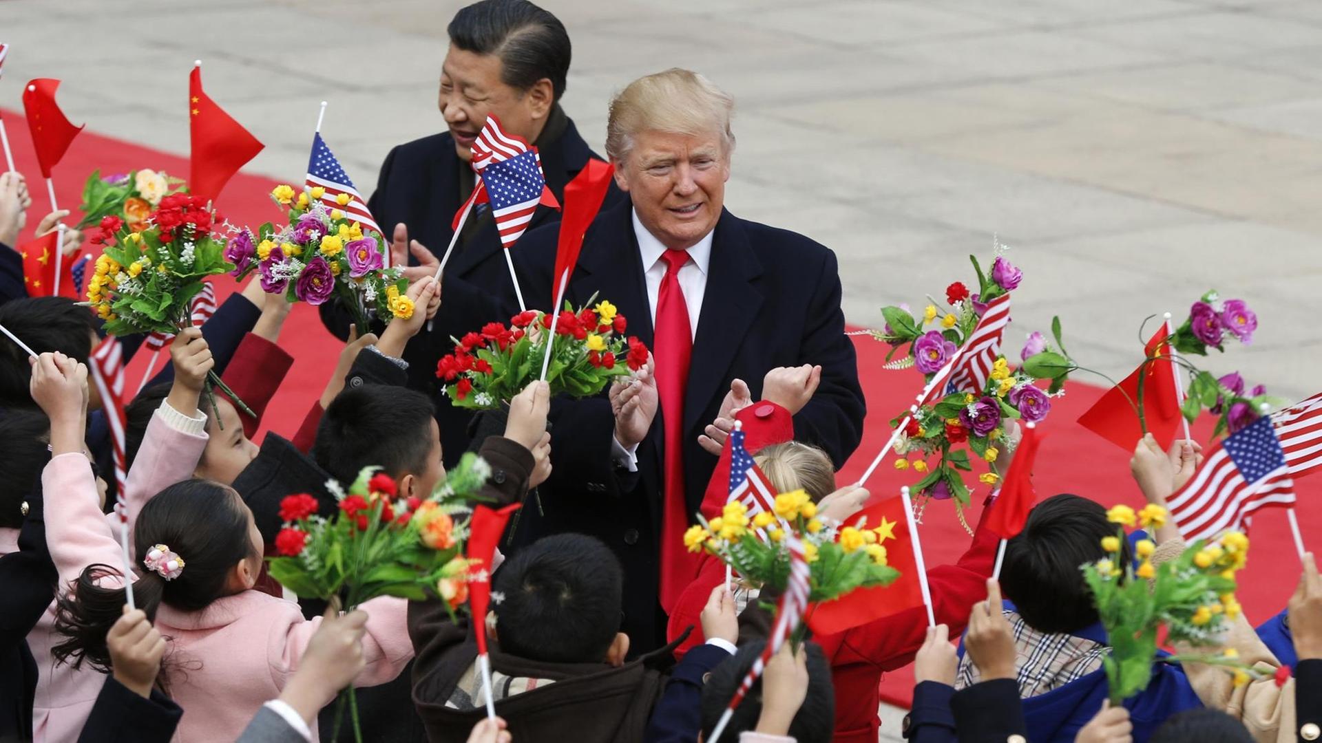 Donald Trump (r.) und Xi Jinping werden von Blumen und Flaggen schwenkenden Kindern begrüßt. Donald Trump (r.) und Xi Jinping werden von Blumen und Flaggen schwenkenden Kindern begrüßt.
