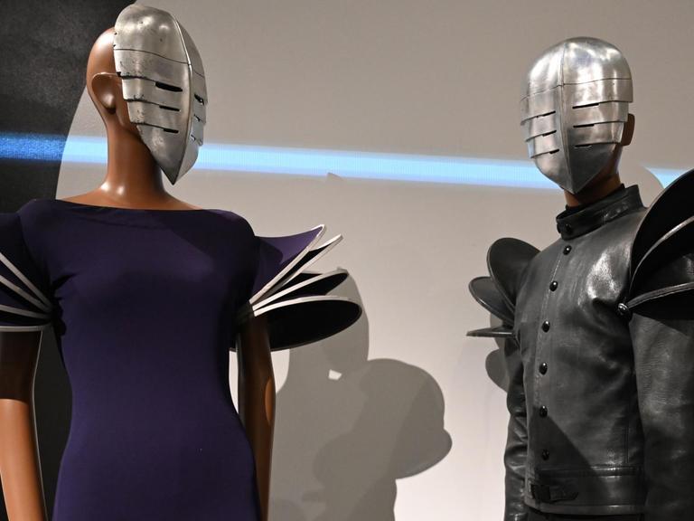 Zwei Schaufensterpuppen in Kleidern von Cardin und mit Masken vor den Gesichtern.