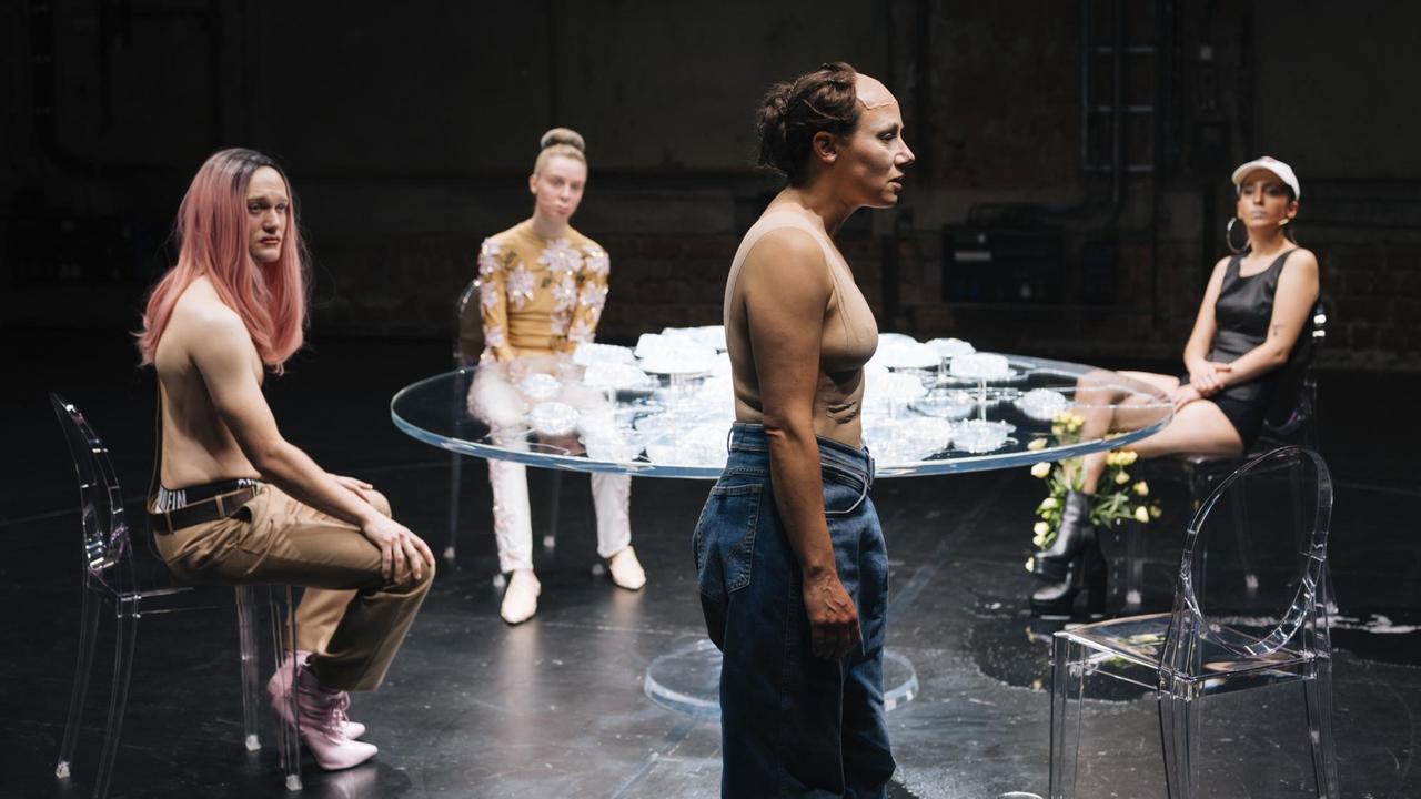 Szenenfoto der Theateraufführung "WUNDE R" in den Münchner Kammerspielen: Drei Menschen sitzen um einen Glastisch und schauen auf eine vierte Person, die davor steht. Auf der Bühne v.l.n.r. die Schauspieler Vincent Redetzki, Julia Windischbauer, Eva Löbau, Zeynep Bozbay