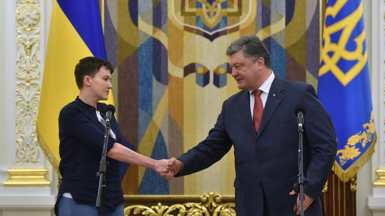 Nadja Sawtschenko und Petro Poroschenko stehen vor Mikrofonen und geben sich die Hand. Im Hintergrund ukrainische Flaggen. Nadja Sawtschenko und Petro Poroschenko stehen vor Mikrofonen und geben sich die Hand. Im Hintergrund ukrainische Flaggen.