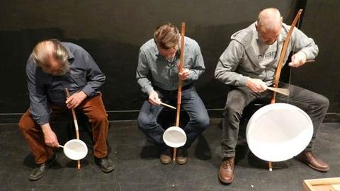 Drei Männer sitzen mit jeweils einem einfach konstruiertem Streichinstrument auf einem Stuhl